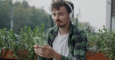 Neşeli Orta Doğulu adam akıllı telefon kullanıyor ve şehir caddesinde oturmuş kulaklıkla müzik dinliyor. Gençlik kültürü ve alet konsepti.