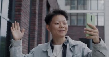 Neşeli Asyalı turist şehir caddesinde yürürken görüntüsünü gösteren ve akıllı telefon ekranına bakan bir video konuşması yapıyor. İletişim ve insan kavramı.