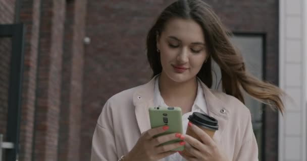 スマートフォンで音声メッセージを録音し コーヒーを飲みながら街角を歩く若い女性のゆっくりとした動き コミュニケーションとモダンなデバイスのコンセプト — ストック動画