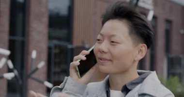 Şehir caddesinde yürüyen ve cep telefonuyla konuşan neşeli Asyalı adamın yakın plan portresi. Modern iletişim ve kentsel gençlik kavramı.