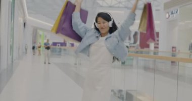 Alışveriş torbalarıyla dans eden neşeli Asyalı öğrenci kulaklıkla müzik dinliyor alışveriş merkezinde eğleniyor. Modern gençlik yaşam tarzı ve tüketim konsepti.