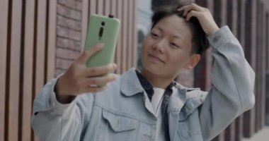 Akıllı telefon kamerasına poz veren neşeli Asyalı adam şehirde selfie çekerken eğleniyor. Milenyum ve aletler kavramı.