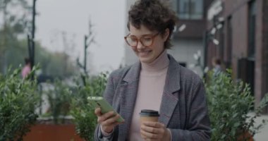 Akıllı telefonuyla mesajlaşan, gülen ve içen bir kız öğrenci kampüste ayakta kahveyi alıp götürüyor. Araçlar ve iletişim konsepti.