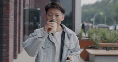 Şen şakrak Asyalı bir adamın akıllı telefon kullanıp kahve içerek caddede yürümesi. Modern gençlik yaşam tarzı ve milenyum konsepti.