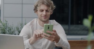 Mobil uygulama kullanan neşeli girişimci elinde akıllı telefon ile sokak kafesinde dizüstü bilgisayarla oturuyor. İletişim ve serbest meslek kavramı.
