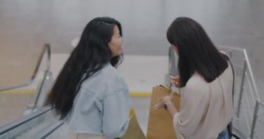 Genç kadınların alışveriş merkezindeki merdivenlerden aşağı inip çantalara bakmaları ve konuşmaları. Boş zaman aktivitesi ve tüketim konsepti.