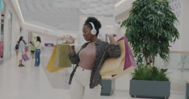 Afro-Amerikan neşeli kadın kulaklık takıyor alışveriş merkezinde alışveriş torbalarıyla dans ediyor ve müzik dinliyor. Moda kıyafetleri içinde mutlu bir bayan mağazada boş vakitlerin tadını çıkarıyor..