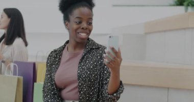 Afro-Amerikalı neşeli bir kadın alışveriş merkezinde akıllı telefonuyla konuşuyor ve gülümsüyor. İletişim ve perakende kavramı.