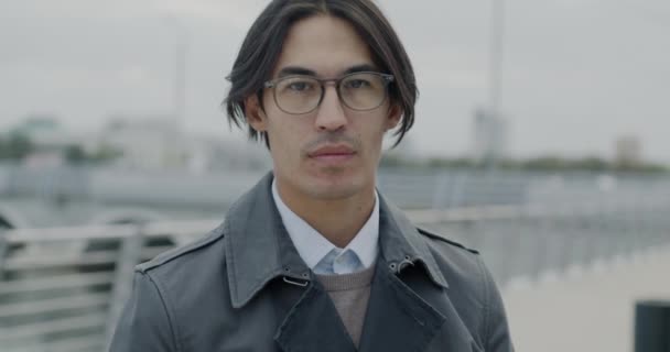 都市に立ってカメラを見ているトレンディな眼鏡を身に着けている自信のあるアジアの起業家のゆっくりとした動きの肖像画 ビジネスマンと都市ライフスタイルのコンセプト — ストック動画