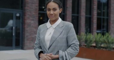 Sokakta durup kameraya bakarak gülümseyen zarif takım elbiseli başarılı genç iş kadınının yavaş çekim portresi. İş ve milenyum konsepti.