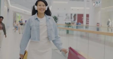 Alışveriş merkezinde alışveriş torbalarıyla yürüyen mutlu Asyalı kadın kulaklıklı müzikle dans edip eğleniyor. Tasasız insanlar ve modern yaşam tarzı konsepti.