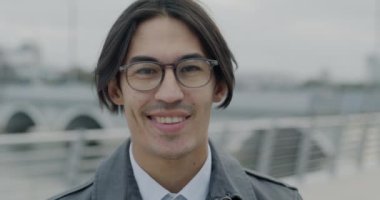 Modern şehir sokaklarında gülümseyen şık gözlükler takan neşeli Asyalı adamın yavaş çekim portresi. Pozitif duygu ve iş adamı konsepti.