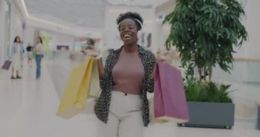 Alışveriş torbaları tutan mutlu Afro-Amerikan öğrenci alışveriş merkezinde kulaklıkla müzik dinliyor. Perakende ve eğlence etkinliği konsepti.