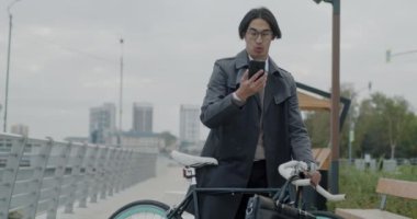 Neşeli Asyalı iş adamı akıllı telefonuyla çevrimiçi video görüşmesi yapıyor. Şehir merkezinde bisikletli duruyor. İnternet iletişimi ve sanal buluşma kavramı.