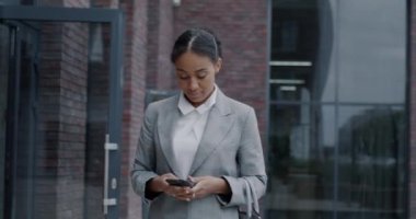 Afro-Amerikalı güzel bir iş kadınının şehir caddesinde yürüyüp akıllı telefon mesajları kullandığı bir fotoğraf. İletişim ve girişimci konsepti.