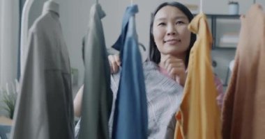 Asyalı güzel bayan elbise asıyor, elbise koleksiyonuna bakıyor, dairede gülümsüyor. Moda ve günlük yaşam tarzı konsepti.