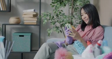 Lastik eldiven takan neşeli Asyalı kadın akıllı telefon içeriğini kontrol ediyor. Daireyi temizledikten sonra kanepede oturup gülüyorlar. Ev işleri ve teknoloji konsepti.