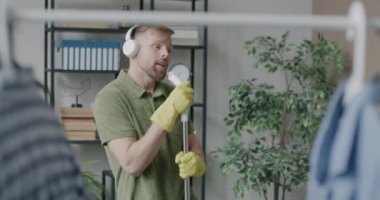 Tek başına dans eden, müzik dinleyen ve paspasla evi temizleyen kaygısız genç adam. Gençlik kültürü ve ev idaresi kavramı.