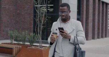 Afro-Amerikalı iş adamı akıllı telefon kullanarak ve kahve içmek için şehir caddesinde yürüyor. Modern yaşam tarzı ve aygıt konsepti.