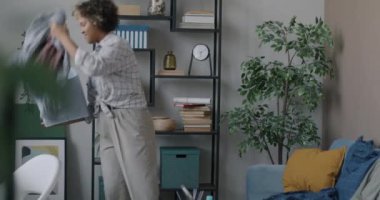 Kaygısız Afrikalı Amerikalı kadın evi temizliyor, eşyaları sınıflandırıyor oturma odasında yürüyor. Düzenli daire ve rutin ev işleri konsepti.