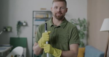 Koruyucu lastik eldiven giyen, elinde paspas tutan ve kameraya gülümseyen genç bir adamın yavaş çekim portresi. Temizlik ve insan konsepti.