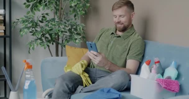 Young Man Using Smartphone Resting Sofa Wearing Rubber Gloves Cleaning — Stock Video