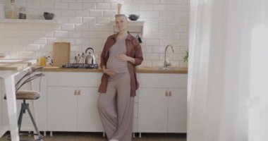 Güzel, şık hamile bir kadının yavaş çekimde çekilmiş portresi. Mutfakta kameraya bakıp gülümsüyor. Hamilelik ve ev hayatı kavramı.