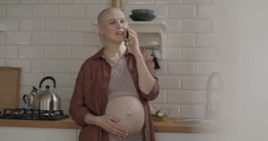 Neşeli genç bir kadın cep telefonuyla gülüyor ve mutfakta konuşmaktan keyif alıyor. İletişim ve hamilelik kavramı.