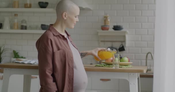 自宅のキッチンでカメラを見ながら笑顔で飲むボトルからオレンジジュースを注ぐ幸せな妊婦のゆっくりとした動きの肖像画 妊娠と健康的なライフスタイルの概念 — ストック動画
