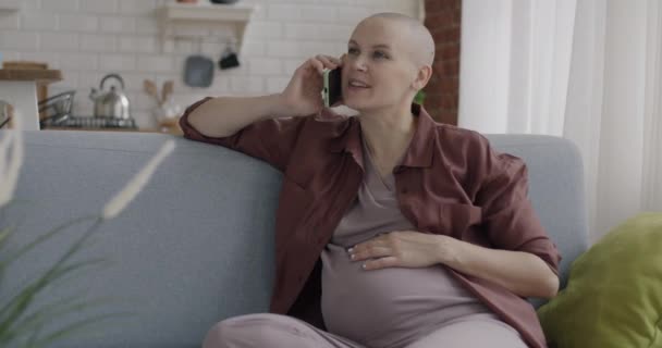 アパートのソファーに座っている腹に触れる携帯電話で話している楽しい妊婦 携帯電話のコミュニケーションと妊娠の概念 — ストック動画