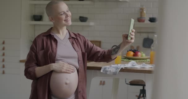 アパートのキッチンで腹に触れるスマートフォンカメラのためのセルフィーを取る陽気な妊婦のゆっくり動き 写真と妊娠コンセプト — ストック動画