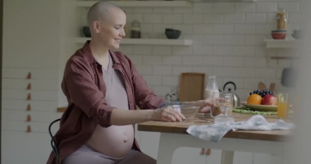 自宅でキッチンで朝食をとるミルクでグラノーラを食べる陽気な妊婦のゆっくりとした動き 健康的な栄養と妊娠の概念 — ストック動画