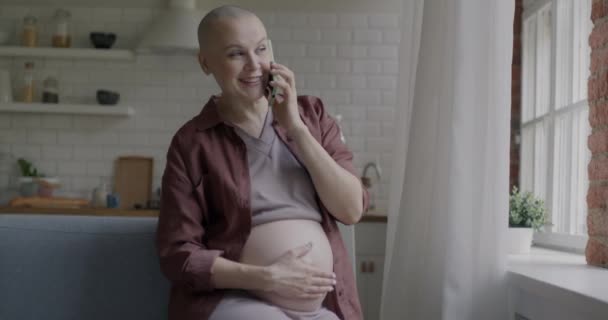 携帯電話で話す楽しい妊婦は 自宅のキッチンに座っている腹に触れながら笑います セルコミュニケーションと妊娠コンセプト — ストック動画
