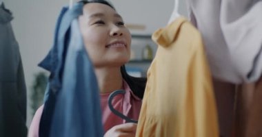 Neşeli Asyalı kadın askısında şık elbiseler asılı moda kıyafet koleksiyonuna bakıyor evde gülümsüyor. İnsanlar ve moda konsepti.