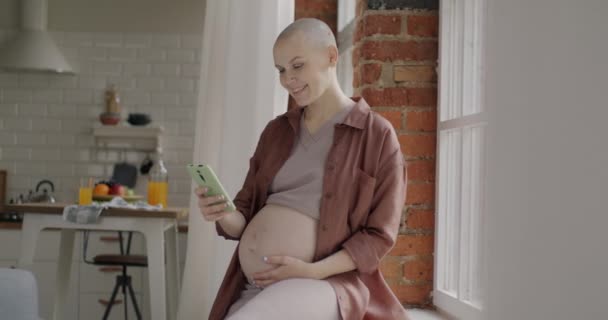 ハッピー妊娠中の女性は 自宅の窓に座ってスマートフォンの笑顔を使用してオンラインコンテンツを楽しんでいます ガジェットと妊娠のコンセプト — ストック動画