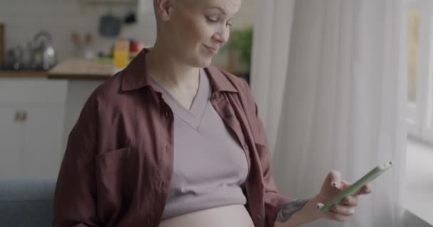 自宅でキッチンに座っているオンラインタッチスクリーンでスマートフォンチャットを使用している妊婦の肖像画 現代のライフスタイルと妊娠のコンセプト — ストック動画
