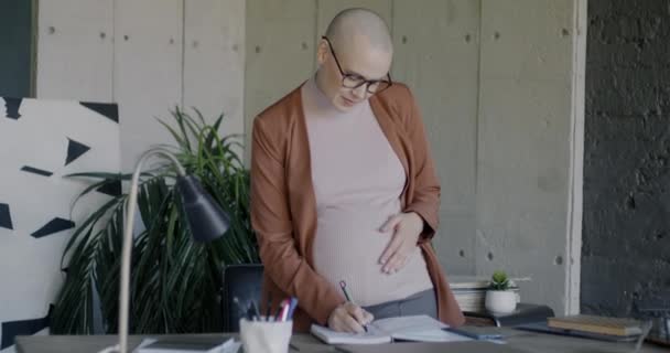 描述怀孕的女商人在笔记本上写字 然后看着相机 微笑着坐在办公室的桌子上 成功的商业和怀孕概念 — 图库视频影像