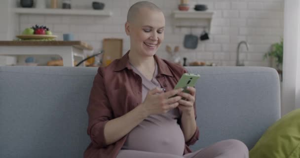 キッチンでソファーに座っているコンテンツを笑顔で共有するスマートフォンでケアフリーの妊婦メッセージ ガジェットとコミュニケーションのコンセプト — ストック動画
