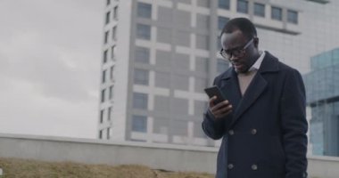 Afro-Amerikan iş adamının yavaş çekimde çekilmiş bir portresi. Şehir caddesinde yürüyor ve akıllı telefon mesajlarını kontrol ediyor. Mobil iletişim ve iş konsepti.