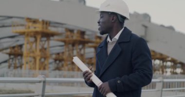 Afrika kökenli Amerikalı mimar koruyucu mimar, inşaat alanında dikilip planlarını tutarken yavaş çekim portresi. İş kurma ve mühendislik kavramı.