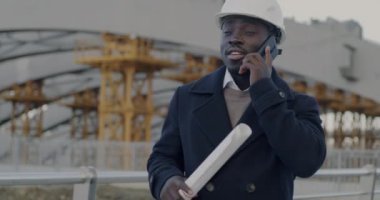 İnşaat alanındaki cep telefonundan konuşan Afrika kökenli Amerikalı inşaat mühendisi. Profesyonel iletişim ve işyeri kavramı.
