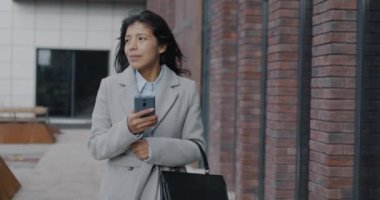 Modern şehirde sokakta yürüyen ve akıllı telefon mesajları kullanan Orta Doğulu genç bir kadının yavaş çekim portresi. İş ve mobil iletişim kavramı.