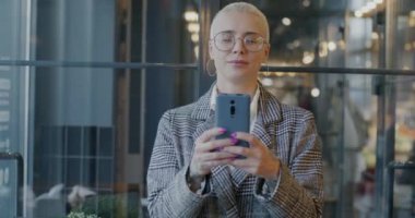 Akıllı telefon kullanan neşeli genç iş kadınının yavaş çekimde portresi sonra kameraya bakıp kafede gülümsüyor. Gadget ve iş konsepti.