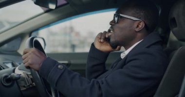 Afro-Amerikan iş adamının şehir caddesinde cep telefonuyla araba sürüşü yavaş çekimde. Modern iletişim ve ulaşım kavramı.