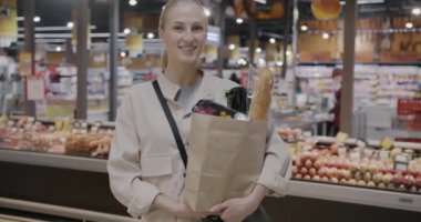 Süpermarkette elinde alışveriş çantasıyla kameraya bakıp gülümseyen neşeli genç bayanın yavaş çekimde portresi. Perakende ve insan konsepti.