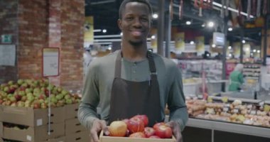 Süpermarkette bir kutu taze elma tutan önlüklü Afrikalı Amerikalı adamın yavaş çekimde portresi. Perakende ve market konsepti.