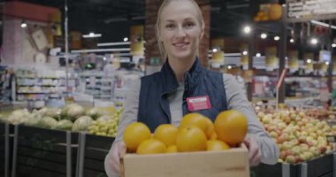 Elinde bir kutu portakal taşıyan ve kameraya gülümseyen genç kadın süpermarket çalışanı portresi. Bakkal ve perakende sektörü kavramı.