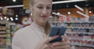 Kulaklık takan, akıllı telefon mesajı kullanan neşeli bir sarışın süpermarkette duruyor. Modern teknoloji ve tüketim kavramı.