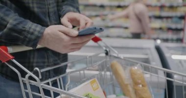 Süpermarkette alışveriş arabası tutan bir adam akıllı telefon kullanırken yakın plan erkek eli. Modern teknoloji ve perakende satış mağazası konsepti.