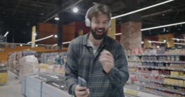 Süpermarkette dans eden, kablosuz kulaklık takan, akıllı telefon kullanan ve dükkanda eğlenen neşeli bir adam. Modern yaşam tarzı ve oyuncu kişi konsepti.